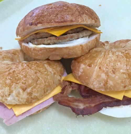 Three breakfast sandwiches on a light surface: one with sausage and cheese, one with ham and cheese, and one with bacon and cheese.