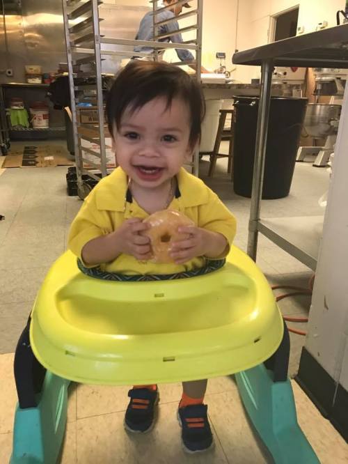 A smiling toddler in a yellow shirt holds a donut while sitting in a high chair in a kitchen setting.