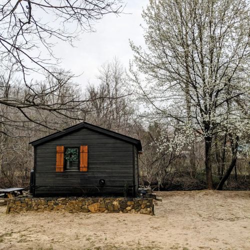 A small black cabin with orange shutters surrounded by trees and a sandy area, with blooming white flowers nearby.