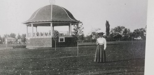 A woman in a long skirt stands in a park near a gazebo on a sunny day, surrounded by trees and grass.