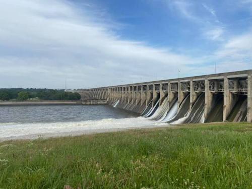 A large dam with water flowing over its spillways, surrounded by green grass and a calm river under a blue sky.