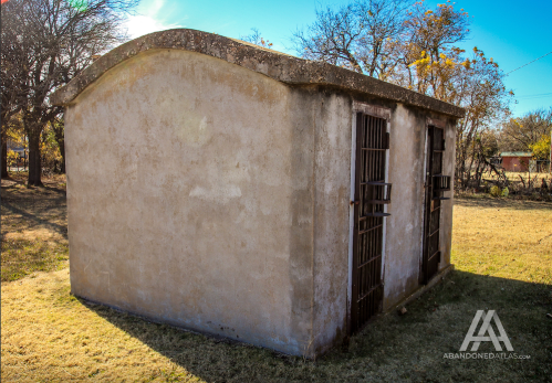 A weathered, concrete structure with barred windows, set in a grassy area surrounded by trees.