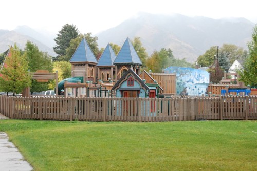 Colorful playground with a castle structure, surrounded by a wooden fence and green grass, set against a misty mountain backdrop.