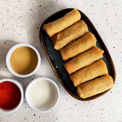 A plate of spring rolls with three dipping sauces: peanut, sweet chili, and white sauce, on a speckled surface.