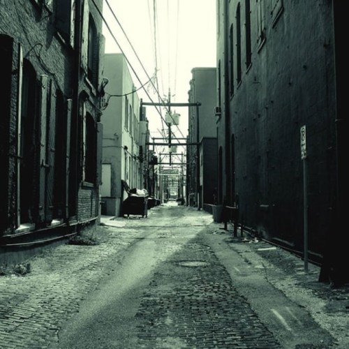 A narrow, dimly lit alleyway lined with brick buildings and power lines, featuring cobblestone pavement.