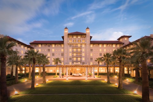 A grand hotel with palm trees in front, illuminated at dusk, showcasing elegant architecture and a welcoming entrance.