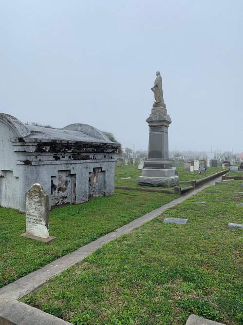 A foggy cemetery scene featuring tombstones and a prominent statue on a pedestal amidst green grass.