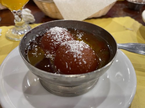 A silver bowl containing two round, syrup-soaked desserts topped with shredded coconut, placed on a white plate.