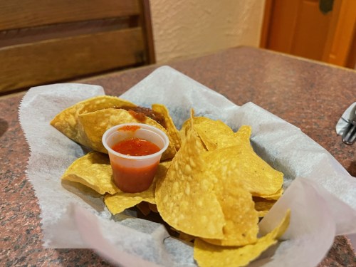 A basket of tortilla chips with a small cup of red salsa on a table.