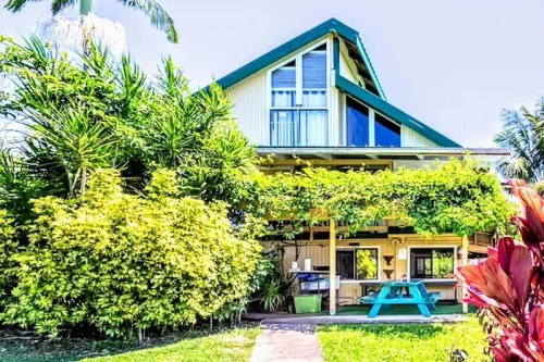 A charming two-story house surrounded by lush greenery and tropical plants, featuring a blue picnic table in front.
