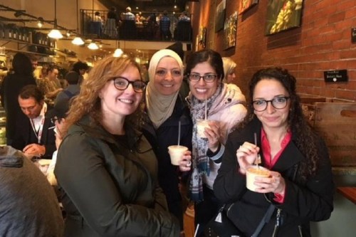 Four women smiling and holding drinks in a busy café, with a warm, inviting atmosphere and brick walls.