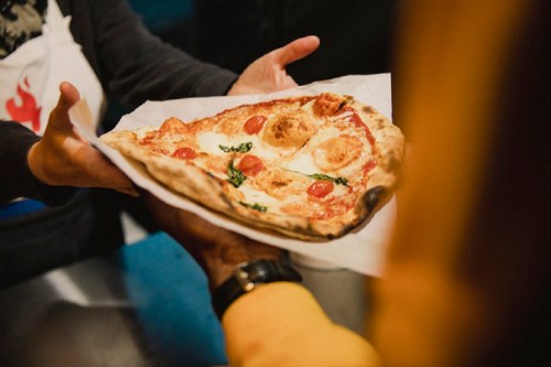 A person hands a freshly baked pizza topped with tomatoes and basil to another person.