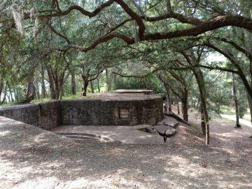 A stone structure surrounded by trees, with a person walking in the distance on a dirt path.