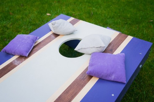 A cornhole game board with a hole in the center and four bean bags in gray and purple colors on a grassy field.