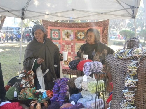 Two women at a craft fair booth, showcasing colorful yarn and handmade items, with a quilt hanging in the background.
