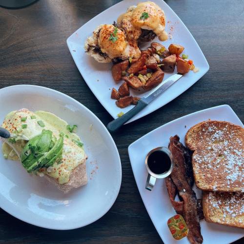 A table with two plates: one with eggs benedict and potatoes, the other with French toast, bacon, and syrup.