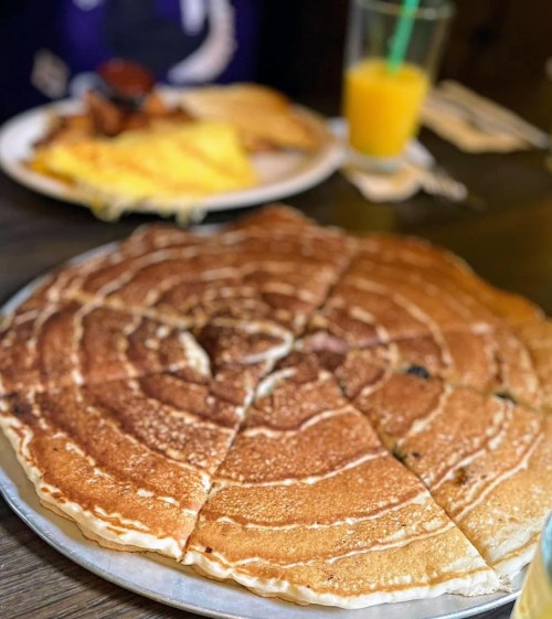 A large, round pancake with a spiral pattern, sliced into pieces, with a plate of eggs and a drink in the background.