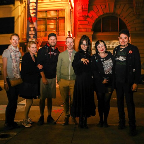 A group of six people poses together at night in front of a building, with festive lighting and a lively atmosphere.
