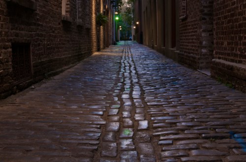 A narrow cobblestone alleyway lined with brick buildings, illuminated by soft green and yellow lights.