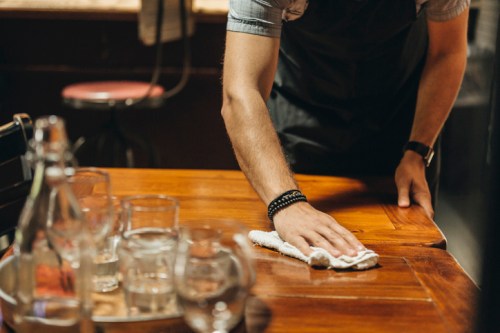 A person in an apron wipes a wooden table with a cloth, surrounded by glasses and a water bottle.