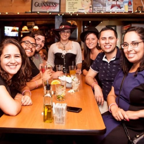 A group of seven friends smiling at a table in a bar, with drinks and a waitress in costume behind them.