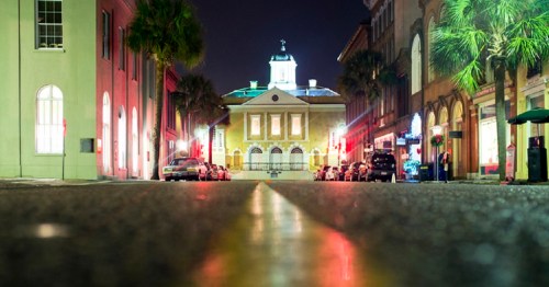 A quiet street at night, lined with palm trees and illuminated buildings, leading to a historic structure in the distance.