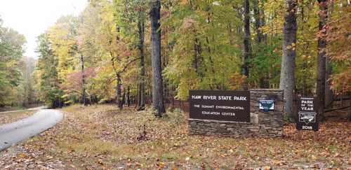 Sign for Haw River State Park with colorful autumn trees lining the path, showcasing nature and educational facilities.