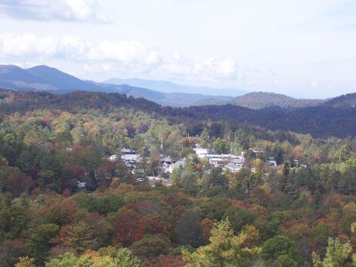 A scenic view of a small town surrounded by colorful autumn foliage and mountains under a partly cloudy sky.