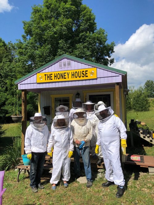 A group of beekeepers in protective gear stands in front of a colorful building labeled "The Honey House."