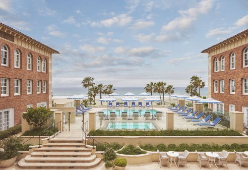 A serene beachfront view featuring a pool surrounded by lounge chairs and palm trees, framed by two brick buildings.