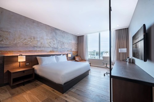 Modern hotel room featuring a large bed, wooden floors, and a large window with city views.