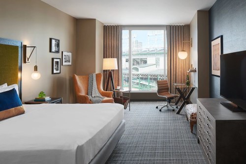 A modern hotel room featuring a bed, armchair, desk, and large window with city views.
