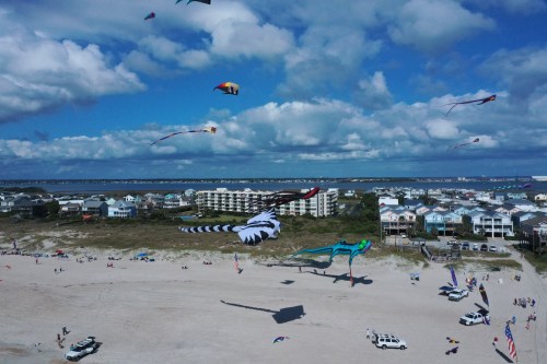 Aerial view of a beach with colorful kites flying above, cars parked nearby, and coastal buildings in the background.