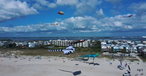 Aerial view of a beach with colorful kites flying over sand and houses, under a blue sky with fluffy clouds.
