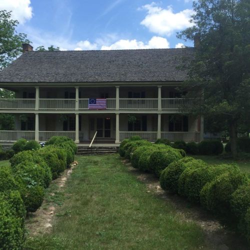 A historic two-story house with a porch, surrounded by neatly trimmed hedges and a blue sky with clouds.