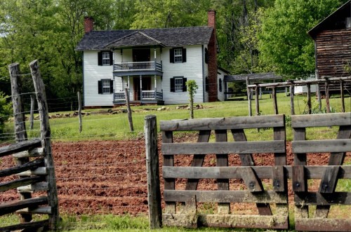 A historic two-story house with a porch, surrounded by green fields and wooden fences.