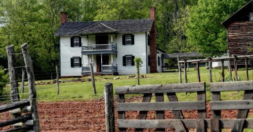 A historic white farmhouse with a porch, surrounded by green trees and a wooden fence, set in a rural landscape.