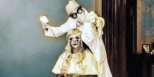 A person in a lab coat and goggles stands behind another person with long blonde hair, both in a spooky setting.