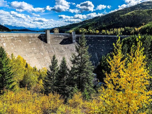 A concrete dam surrounded by autumn trees and mountains under a blue sky with fluffy clouds.