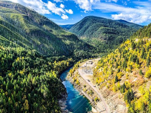 A scenic view of a river winding through lush green mountains, with autumn foliage and a power station visible.