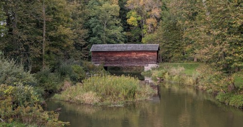 A rustic wooden bridge over a calm stream, surrounded by lush green trees and autumn foliage.