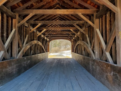 View from inside a wooden covered bridge, showcasing the rustic beams and a glimpse of the landscape beyond.
