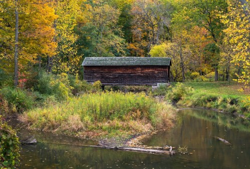 A rustic wooden cabin surrounded by vibrant autumn foliage and a calm stream.