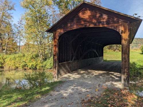 A wooden covered bridge over a calm stream, surrounded by trees with autumn foliage.