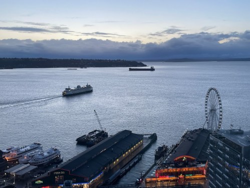 A view of a harbor at dusk, featuring a ferry, a large wheel, and illuminated buildings along the waterfront.