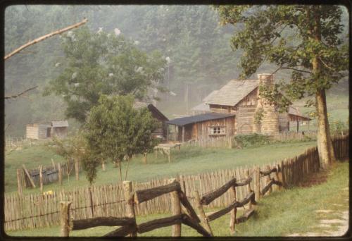 A rustic farmhouse surrounded by trees and a wooden fence, set in a misty landscape.