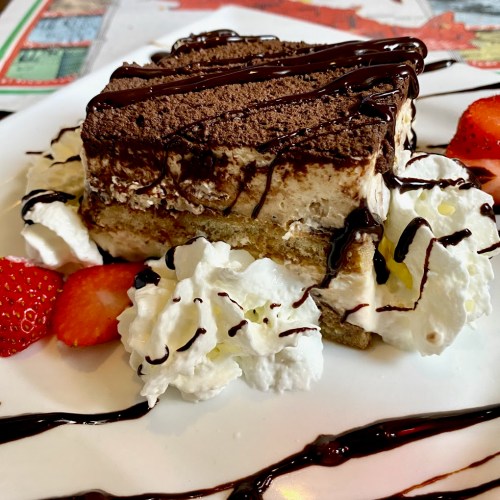 A slice of tiramisu topped with chocolate drizzle, whipped cream, and fresh strawberries on a white plate.