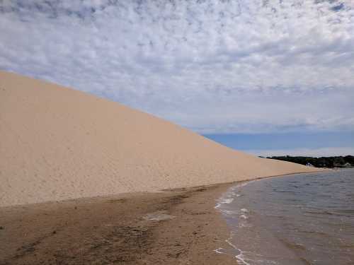 A sandy dune rises beside a calm body of water under a cloudy sky.