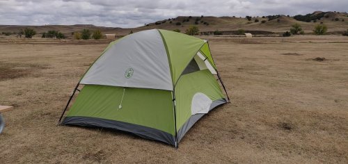 A green and gray tent set up on dry, grassy terrain with a cloudy sky in the background.