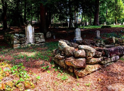A serene graveyard with weathered tombstones surrounded by trees and a stone wall, covered in pine needles and grass.